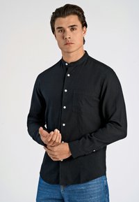 Camicia nera con colletto realizzata in tessuto leggero, caratterizzata da una chiusura a bottoni, maniche lunghe e una tasca sul petto. Indossata con jeans blu.