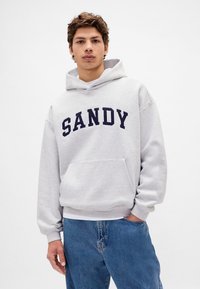 Szara bluza z kapturem z napisem "SANDY" w granatowym kolorze, z kieszenią kangurka i luźnym krojem. Noszona z niebieskimi dżinsami.
