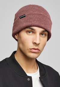 Pletená čepice v burgundské barvě s texturovaným žebrovaným designem. Má černou nášivku s nápisem "URBAN CLASSICS." Nosená modelem s černou bundou.