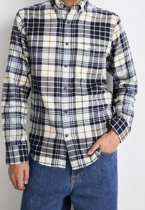 Homme portant une chemise à carreaux bleu marine, blanche, verte et beige, boutonnée, avec une poche poitrine, rentrée dans un jean bleu.