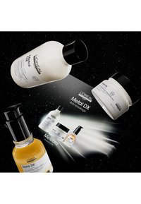 Sett med L'Oréal Metal DX hårpleieprodukter som inkluderer en hvit flaske, en kremkrukke og et gult serum, alt mot en mørk, stjernespekket bakgrunn.