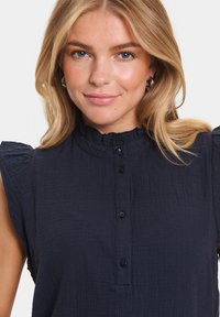 Top fără mâneci de culoare albastru marin, dintr-un material texturat, cu guler volănat și închidere cu butoane. Prezintă un design moale și fluid, cu mâneci puțin bufante.