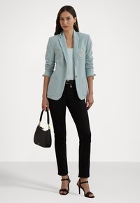 Modra blazer z okrasnim grbom na levem žepu, v kombinaciji s svetlo majico, črnimi slim jeans hlačami in črnimi sandali s trakci.