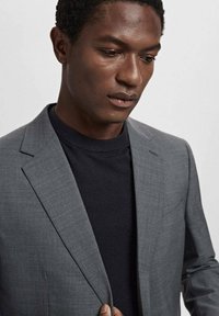 Blazer gris ajusté avec une texture subtile, associé à un col rond en tricot foncé. Comprend des revers à cran et une fermeture à bouton unique.