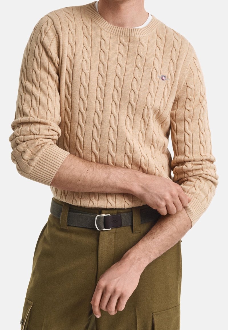 Homme ajustant la manche d'un pull en maille torsadée beige, portant un pantalon vert olive avec une ceinture noire, sur fond blanc.