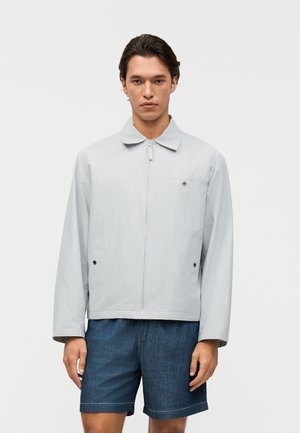 Άνδρας που φοράει ανοιχτό γκρι ζακέτα με φερμουάρ και γιακά και μπλε τζιν σορτς, στέκεται μπροστά σε απλό λευκό φόντο.