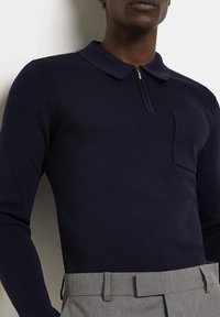 Polo à manches longues bleu marine avec col zippé, détail de poche avant, texture côtelée, associé à un pantalon gris ajusté.