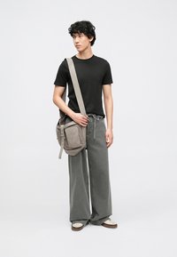 Siva torbica za čez ramo z teksturirano površino in nastavljivim trakom. Model nosi črno majico, široke sive hlače in bež čevlje.