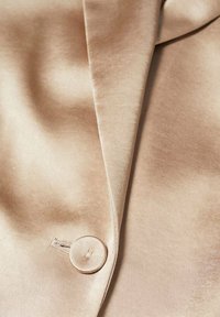 Béžová saténová látka s hladkou texturou, vybavená zapínáním na jeden knoflík a klopami. Jemný lesk a splývavost jsou viditelné.