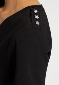 Top negro con escote en V, que presenta tres apliques de estrellas acentuadas con cristales en el hombro. Fabricado con una tela ligera y suave.