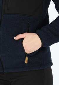 Navy fleece jacket with black accents, featuring a zippered pocket and a textured fabric. Elastic cuffs and hem provide a snug fit.