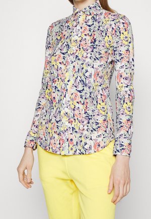 Femme portant une chemise boutonnée à manches longues avec un motif floral multicolore et un pantalon jaune vif.