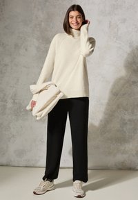 Cremfarbener Rollkragenpullover mit gerippten Bündchen und Saum, kombiniert mit schwarzen, weit geschnittenen Hosen. Trägt eine weiche, crèmefarbene, strukturierte Tasche. Legere Schuhe.