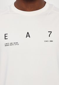 Balta sportinė marškinėliai su juodu tekstu: "EA 7," "Ribos Lūžta Su Stiliumi," ir "Nuo 2004." Lygi tekstūra, apvalus kaklas.