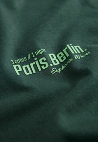 Dunkelgrüner Stoff mit weißem Text, der "3 Städte // 1 Nacht" und "Paris.Berlin." liest, mit "Euphoria Waves" in einer helleren Farbe.