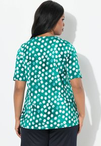 Teal T-shirt met witte stippen, korte mouwen, losvallende pasvorm, gladde stof en gebogen zoom, gezien vanuit de rug.