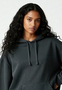 Giovane donna con lunghi capelli neri ricci, che indossa un ampio felpa scura, che guarda direttamente verso la macchina fotografica con un'espressione neutra.