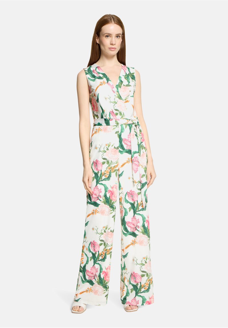 Femme portant une combinaison blanche sans manches imprimée de fleurs roses et vertes avec une ceinture assortie, debout devant un fond uni.