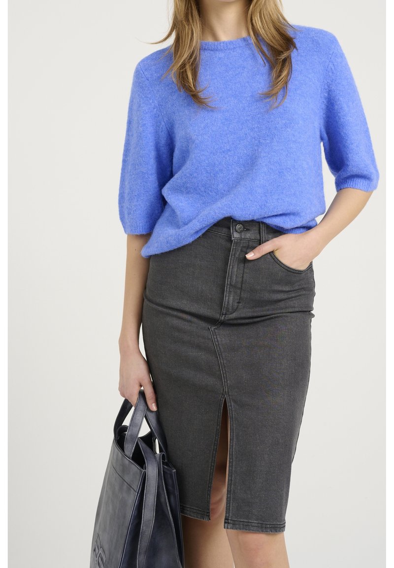 Pull en maille duveteuse bleu clair à manches courtes associé à une jupe crayon en denim foncé avec une fente frontale. Le mannequin tient un sac fourre-tout foncé.