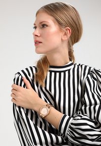 Reloj de oro rosa con esfera redonda, acentos de cristal y correa de metal, llevado en una muñeca con una blusa de mangas abullonadas a rayas en blanco y negro.