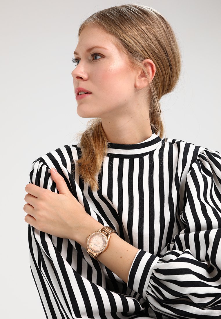 Reloj de oro rosa con esfera redonda, acentos de cristal y correa de metal, llevado en una muñeca con una blusa de mangas abullonadas a rayas en blanco y negro.