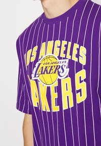 Camiseta a rayas moradas con el logo de "LOS ANGELES LAKERS" y un gráfico de baloncesto. Diseñada para los aficionados de los Lakers, enfatizando el espíritu de equipo.