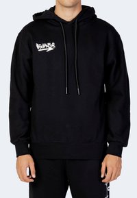 Uomo che indossa un hoodie nero con la scritta bianca "Disclaimer" e una freccia sul petto, abbinato a pantaloni neri che mostrano un parziale design bianco.