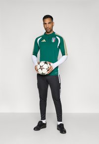 Πράσινη μπιγκόν Adidas με λευκές λεπτομέρειες και λογότυπο Ιταλίας, σε συνδυασμό με μαύρα παντελόνια που έχουν λευκές ρίγες και μαύρα παπούτσια ποδοσφαίρου, κρατώντας μια μπάλα.