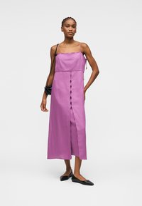 Femme debout portant une robe midi sans manches violette avec une fente à l'avant et des ballerines noires pointues, tenant un petit sac à bandoulière noir.