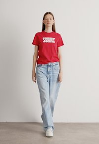 Punane puuvillane T-särk, millel on valge "TOMMY JEANS" tekst, koos helesiniste, lõdvalt istuvate teksade ja valgete tennisadenä.