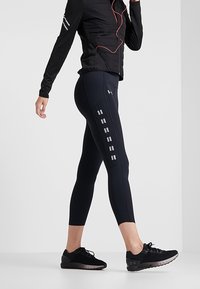 Černé sportovní legíny s reflexními akcenty a přiléhavým střihem, doplněné černou zipovou bundou a černými běžeckými botami.