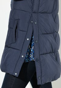 Marinblå pufferväst med ett quiltat mönster, tryckknappar och sidofickor, lagd över en blåmönstrad skjorta och mörka jeans.