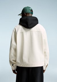 Cream-colored jacket with a black hood, featuring a textured fabric, relaxed fit, and side vents, paired with a green cap.