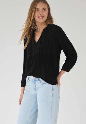 Kvinde iført en sort bluse med lange ærmer med broderede detaljer og lyseblå højttaljede jeans, stående med den ene hånd i lommen.