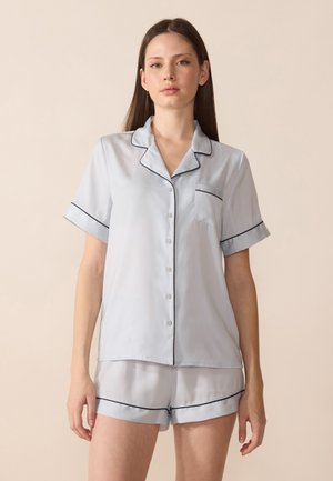 Light blue silk pajama set featuring a short-sleeve button-up top with a pocket and black piping, paired with matching shorts.