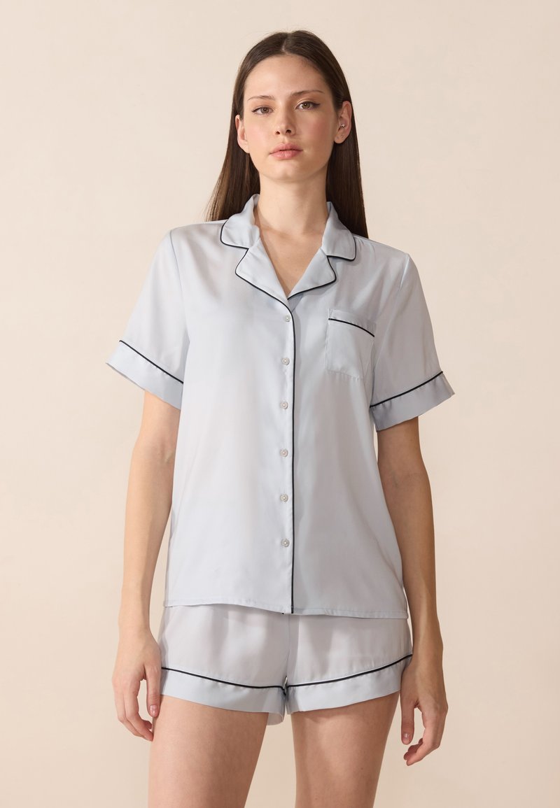 Set di pigiama in seta azzurro chiaro composto da una camicia a maniche corte con bottoni, una tasca e piping nero, abbinato a pantaloni corti coordinati.