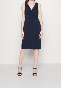 Marinblå ärmlös klänning med V-ringning, omslagdesign och sned bälte detaljer. Slät tyg med knälång längd.