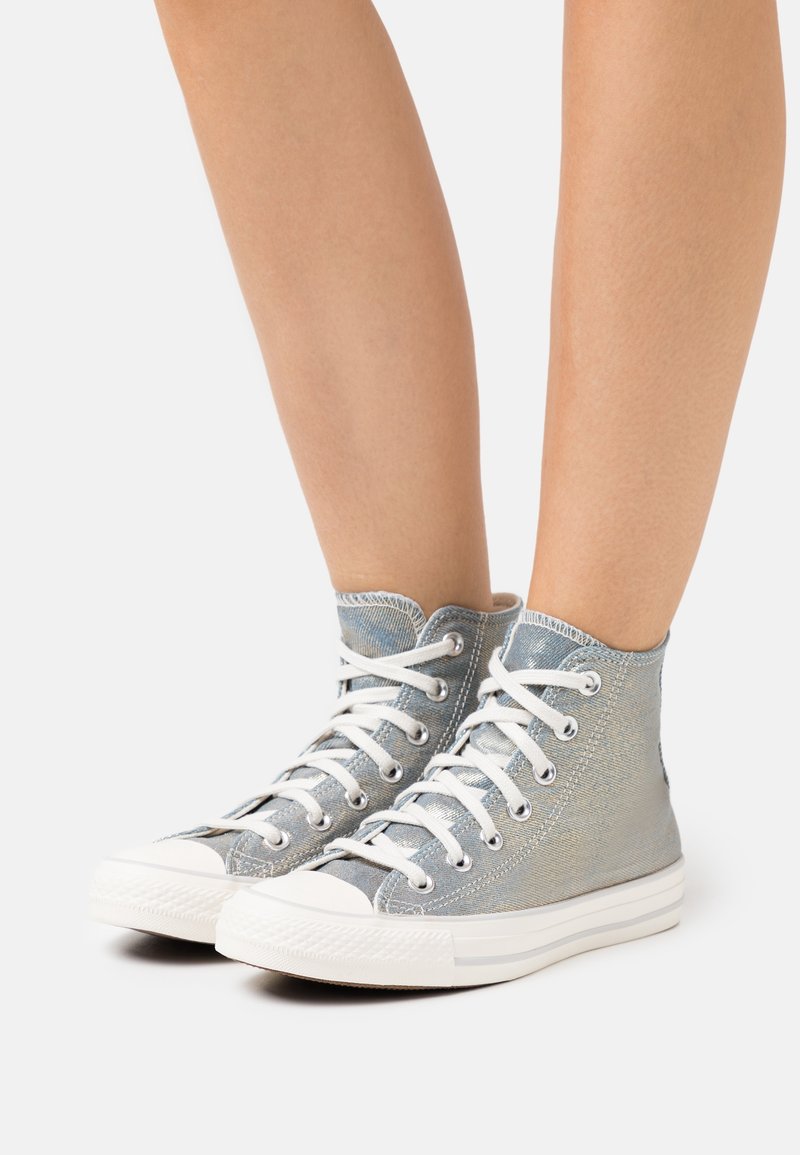 Jambes inférieures portant des baskets montantes en toile argentée avec des lacets blancs et des semelles en caoutchouc sur un fond blanc.