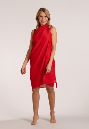 Woman standing barefoot, smiling, wearing a sleeveless red knee-length dress with a draped design against a plain light background.