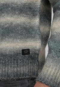Suéter de punto con un degradado de tonalidades gris y crema, con una textura suave y puños acanalados. Incluye un parche de marca negro cerca del dobladillo.