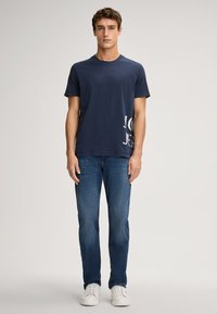 Navyfarbener Baumwoll-T-Shirt mit Rundhalsausschnitt und weißem Grafiktext an der Seite, kombiniert mit blauen Jeans und weißen Sneakers.