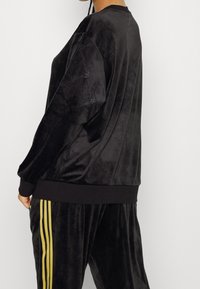 Osoba oblečená v černém sametovém teplákovém kompletu s embosovanými vzory na vršku a kalhotami se zlatými pruhy, stojící na jednobarevném pozadí.