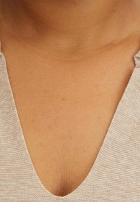 Ljus beige stickad topp med en djup V-ringning, som visar en texturerad väv. Synlig hudton under ringningen.