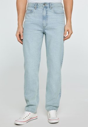 Hele sinised sirge säärega teksapüksid, mis on kantud valgete madalate tennistega, nähtavad vööst jalgadeni ühevärvilisel taustal.