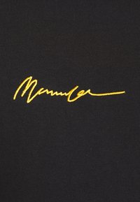 Tessuto nero con una superficie liscia che presenta un evidente design firmato ricamato in giallo. I punti di cucitura contrastano con il colore di sfondo.