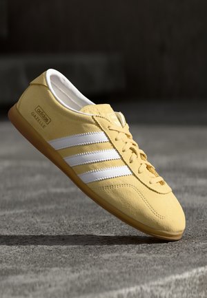 Żółty but sportowy Adidas Gazelle z białymi paskami i gumową podeszwą, ustawiony pod kątem nad szarą betonową powierzchnią.
