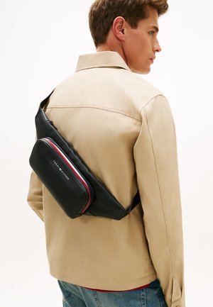 Melnā plecu soma ar kontrastējošiem sarkaniem un baltiem akcentiem, ko nēsā cilvēks ar bēšas krāsas jaku un džinsa biksēm.