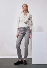Szürke farmernadrág elülső zsebekkel és slim fit szabással, fehér, hosszúujjú toppal párosítva, világos bézs kötött sállal rétegezve. Fekete cipő.
