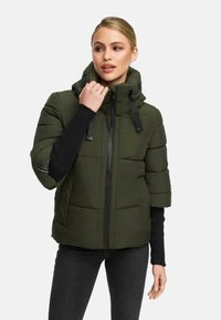 Woman wearing an olive green puffer jacket with short sleeves over a black long-sleeve top and black pants, adjusting the jacket collar.