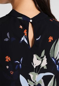 Blouse noire à motifs floraux avec une ouverture en forme de goutte boutonnée au dos, présentant un mélange de motifs floraux verts, bleus et orange sur un tissu lisse.